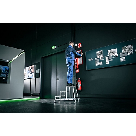 Un homme en bleu de travail est debout sur un escabeau et attache quelque chose à un mur. En arrière-plan, un mur noir avec une chronologie.