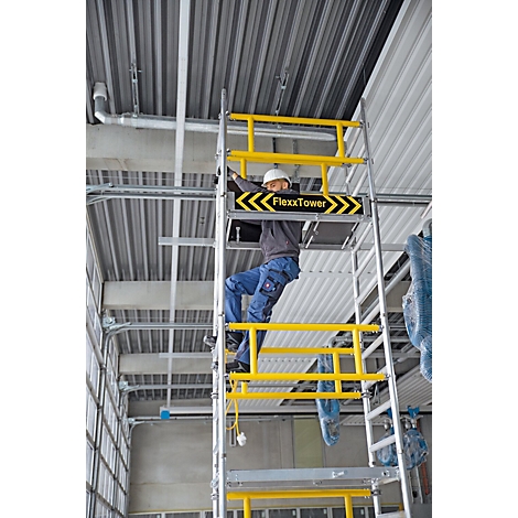 Ein Mann auf einem Gerüst montiert etwas an der Decke. Das Gerüst hat gelbe Sicherheitsgeländer. Auf dem Gerüst steht "FlexxTower".