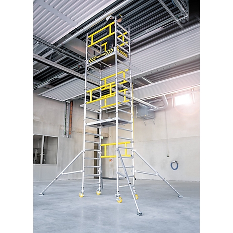 Een steiger met gele veiligheidsleuningen staat in een industrieel gebouw. Bovenin is een persoon te zien.