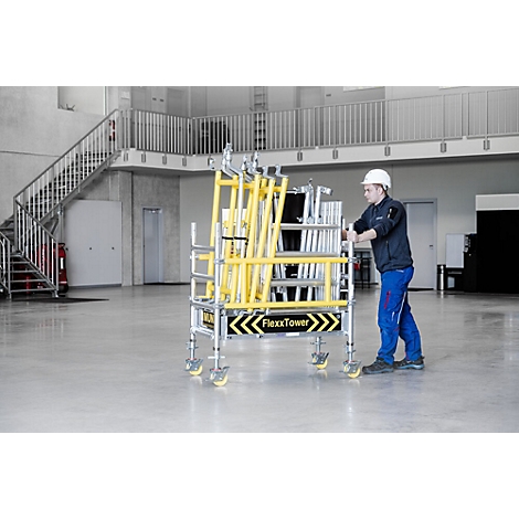 A man pushing a yellow FlexxTower scaffolding cart on wheels in a warehouse. A staircase in the background.
