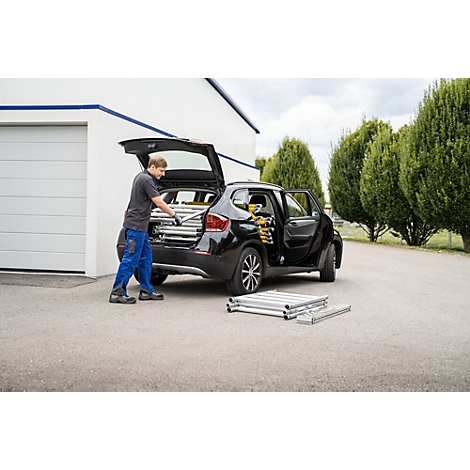 Een man laadt ladders in een auto. Open achterklep, een gereedschap in de open deur, bomen op de achtergrond.