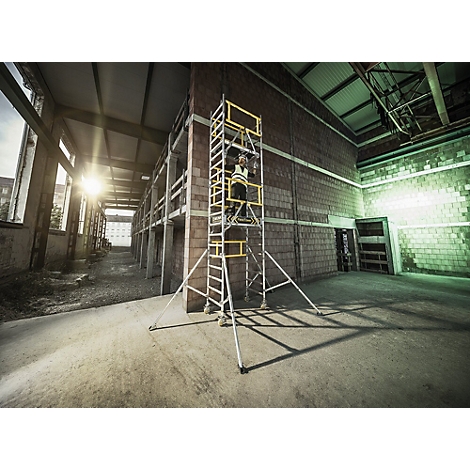Persoon op steiger voor bakstenen muur. Werkkleding, gele details, industriehal.