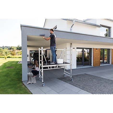 Man paints the outside wall with a roller, woman paints on the side. Both are standing on a scaffolding platform.