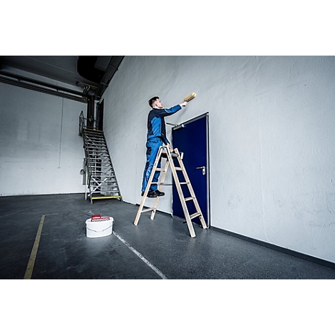 Een man in blauwe werkkleding schildert een muur. Hij staat op een ladder, met een blauwe deur achter hem.