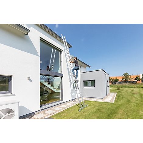 Een persoon beklimt een ladder tegen een witte huismuur. Een grijs bijgebouw en gazon zijn ook zichtbaar.