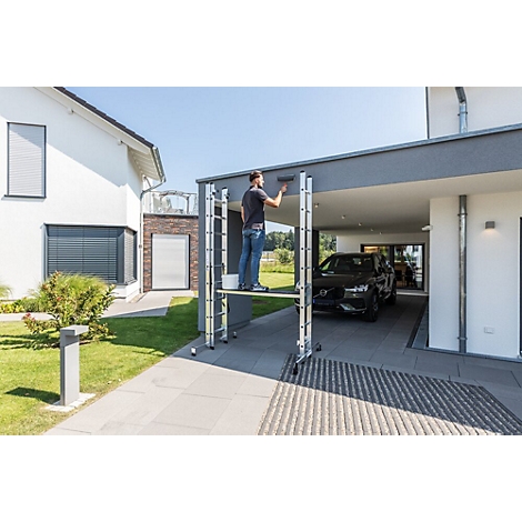 Man schildert de muur van het huis, staand op een steiger. Op de achtergrond een garage met een auto en een huis.