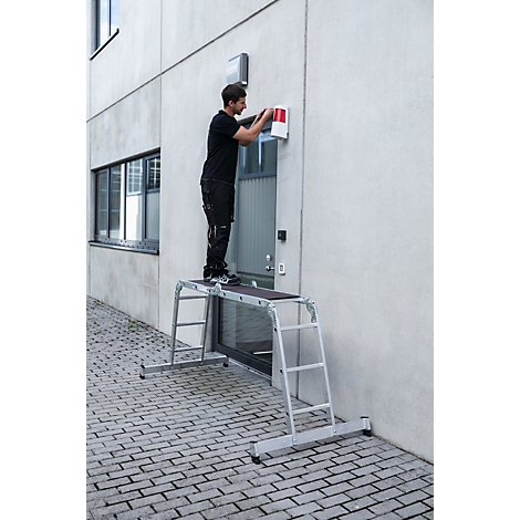 Een man werkt aan een muur, staand op een werkplatform. De muur is wit en heeft een deur en een raam.
