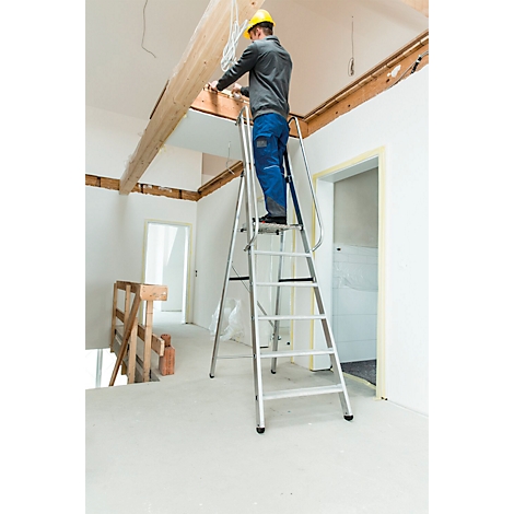 Ein Handwerker auf einer Leiter montiert Kabel an einer Decke. Das Innere eines Hauses ist im Bau. Es gibt Holzbalken und weisse Wände.