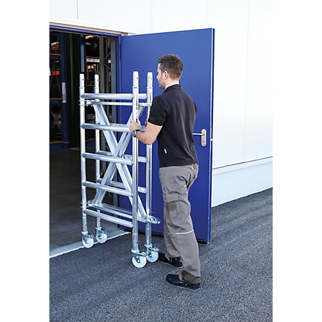 A man pushes a metal scaffolding through a blue door. Scaffolding with rollers, gray trousers and black shirt.