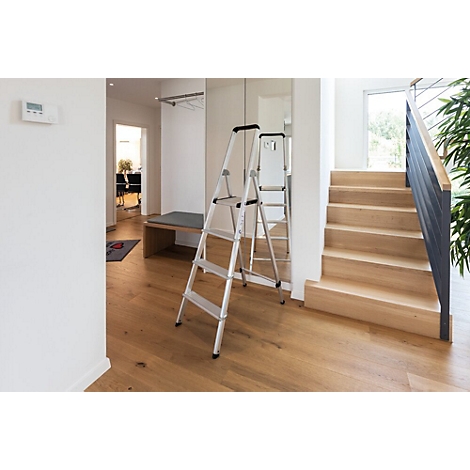 In einem Flur steht eine silberne Stehleiter. Rechts eine Holztreppe, links eine Bank und ein Spiegel.