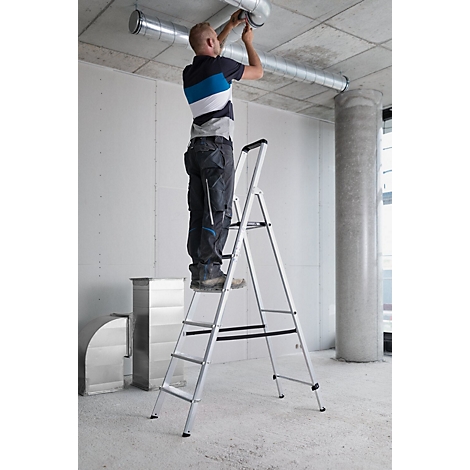 Ein Mann auf einer Leiter arbeitet an einem Rohrsystem. Er trägt Arbeitskleidung. Grauer Betonboden und Decke.