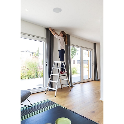 Eine Frau auf einer Leiter befestigt Gardinen in einem hellen Wohnzimmer.