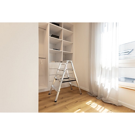 Ein silberfarbener Trittschemel steht vor einem weißen Einbauschrank. Rechts ein Fenster mit Vorhängen.