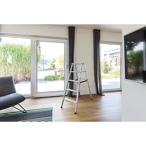 Een kleine ladder staat in een moderne woonkamer. Grote ramen bieden uitzicht op de tuin. Een televisie hangt aan de muur.