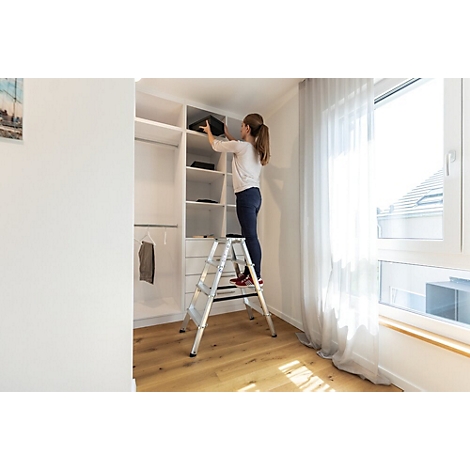 Een vrouw sorteert in een witte kast, staand op een zilveren trap. Lichte kamer met raam.