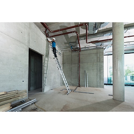 Een bouwvakker op een ladder installeert pijpen in een ruwbouw. Plafonds, muren en vloeren zijn van beton.