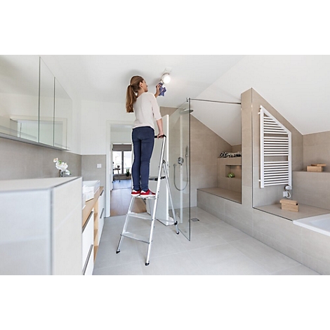 Vrouw staat op een ladder en maakt een plafondlamp schoon in een moderne badkamer.