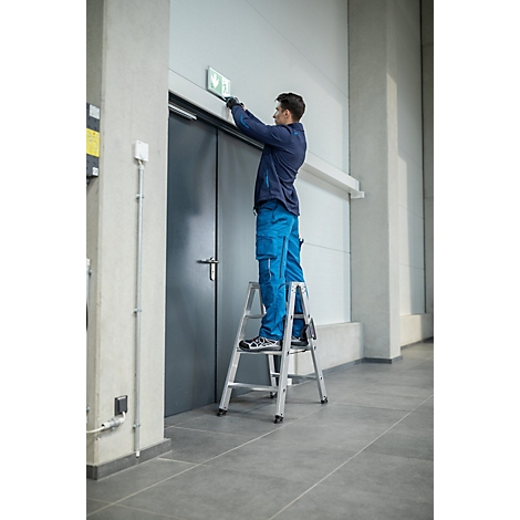 Een man op een ladder bevestigt een nooduitgangsbordje aan een deur.