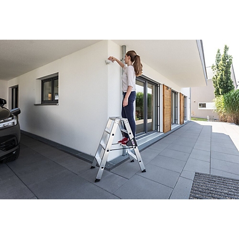 Vrouw op een ladder die een camera aan de muur van het huis bevestigt. Wit huis met grijze grond. De blik van de vrouw is gericht op de camera.