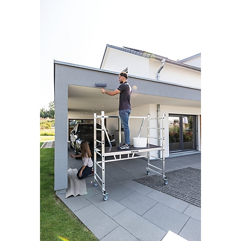 Een man schildert het plafond van een garage, terwijl een meisje op de grond zit. Een ladder staat klaar.