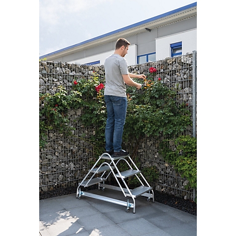 Man staat op een trap, rozen snijdend voor stenen schanskorven. Huis op de achtergrond.