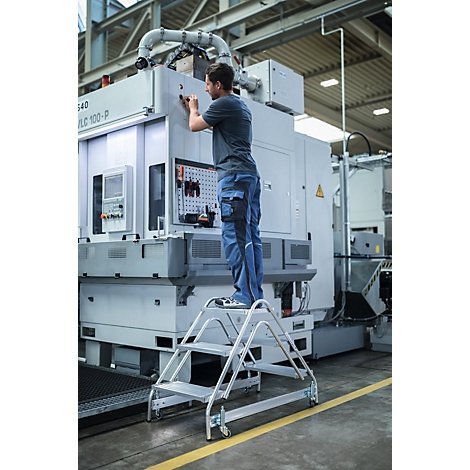 Man op een trap monteert machine in een fabriek. Grijze trap, blauwe broek.