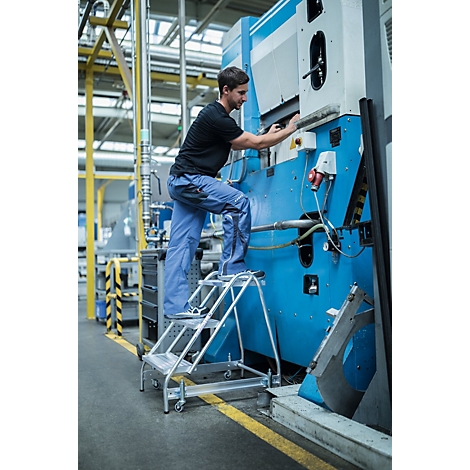 Een man staat op een trap en werkt aan een grote blauwe machine in een fabriek.