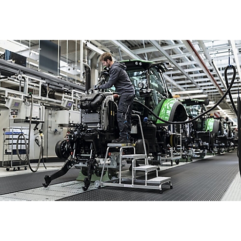 Een man werkt aan een tractor in een fabriek, staand op een kleine trap.