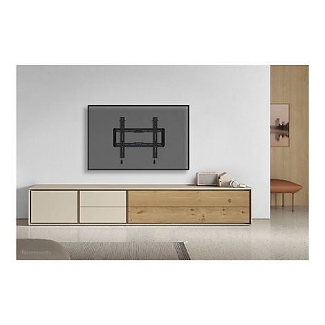 Ein TV-Wandhalter, montiert an einer grauen Wand, über einer modernen Kommode, mit Dekor. Die Marke Neomounts ist unten links zu sehen.