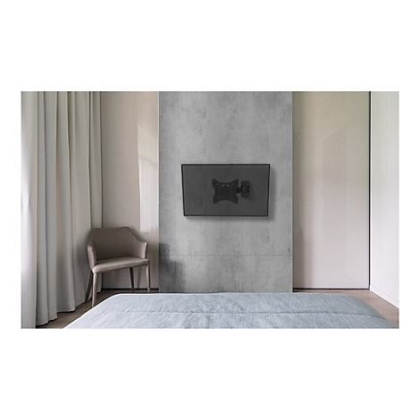 Ein Fernseher hängt an einer grauen Wand über einem Bett. Daneben ein Stuhl und Vorhänge.