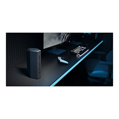 Ein schwarzer Router steht auf einem Schreibtisch. Im Hintergrund: PC-Monitor, Tastatur, Maus und Gaming-Stuhl.