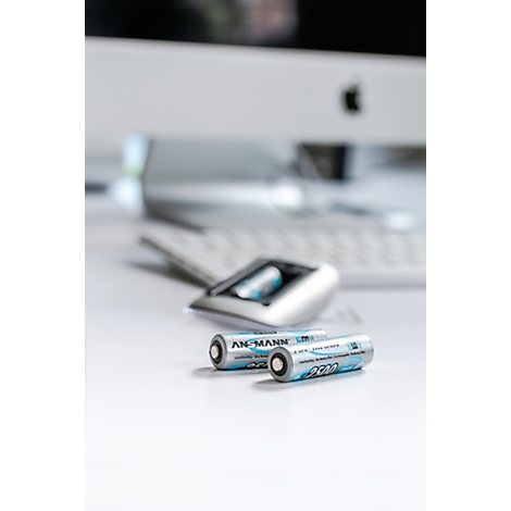 Two batteries with "ANSMANN" print lie in front of a computer. In the background a mouse charger with another battery is visible.