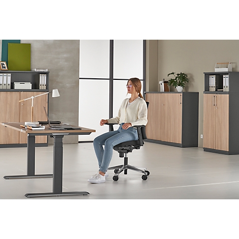 Eine Frau sitzt an einem Schreibtisch in einem Büro, trägt Brille und Jeans. In der Umgebung sind Schränke und Regale.
