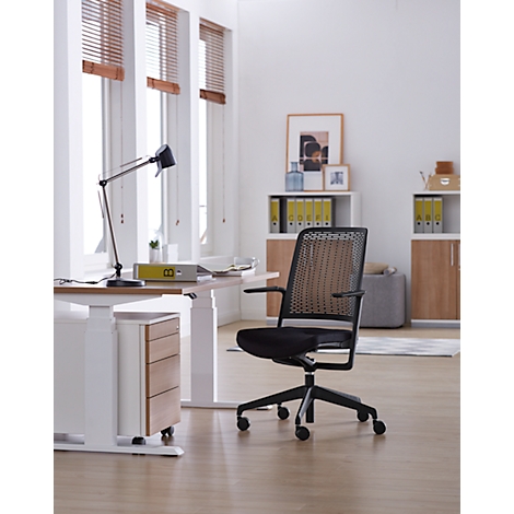 Bureaustoel voor een raam. De stoel heeft een zwart frame en een geweven rugleuning. Het bureau is wit met een houten afwerking.