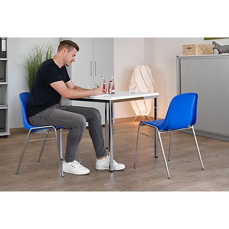 Ein Mann sitzt an einem Tisch, schreibt. Blauer Stuhl, Wasserflasche, Lampe im Hintergrund.