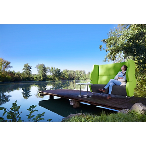 Eine Frau sitzt entspannt auf einem grünen Sofa am Steg eines Sees. Sie genießt die Sonne und hält ein Buch.