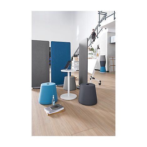 Kantooropstelling met blauwe, grijze en witte elementen. Krukken, scheidingswanden en een tafel met een laptop staan op de houten vloer. Een persoon loopt de trap op.