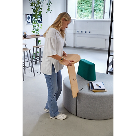 Eine Frau hält einen Holzstuhl. Sie steht in einem Büro. Auf einem runden Sofa liegen Notizbücher und Stifte.