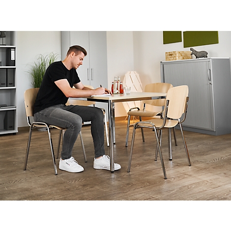 Ein Mann sitzt an einem Tisch und schreibt. Drei weitere Stühle stehen um den Tisch herum. Im Hintergrund ein Schrank.