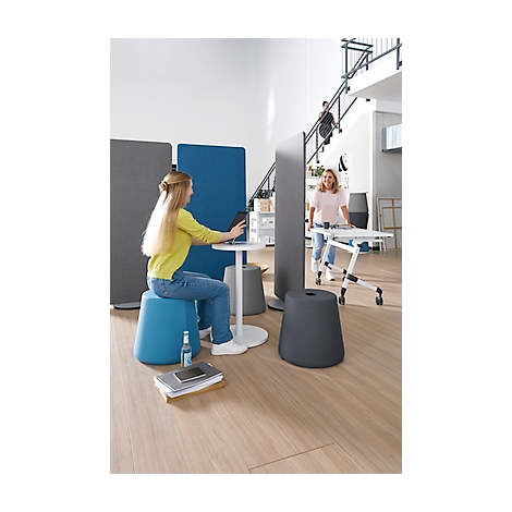 Young woman sitting on a blue stool, working on a tablet. Other people and office furniture in the background.