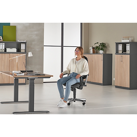 A woman sits in an office chair at a desk. She is wearing jeans, a cream-colored sweater and glasses. Office furniture in the background.