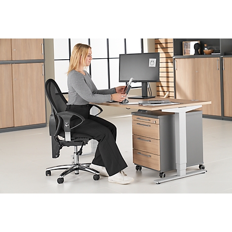 A woman sits at a height-adjustable desk working with a tablet. Next to her is an office chair.