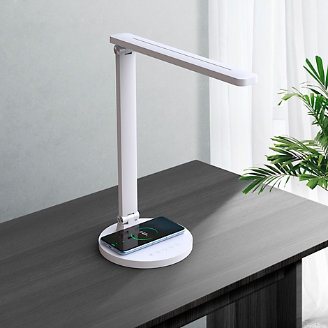 Witte bureaulamp met geïntegreerd oplaadstation met een smartphone erop. De lamp staat op een donker bureau.
