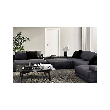 Ein weiÃŸer Projektor auf einem Couchtisch in einem Wohnzimmer mit Eckcouch, Pflanze und Lampe.