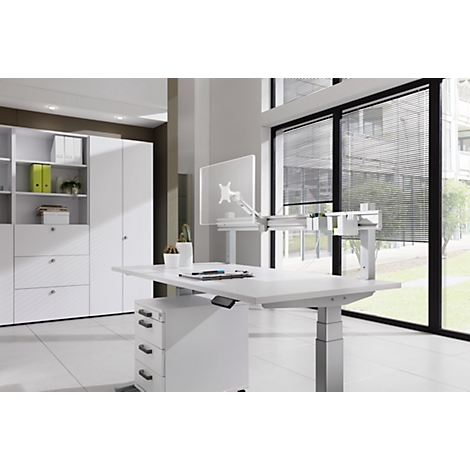 White height-adjustable desk in a bright office. Pens and a monitor are on the table. In the background is a wall unit and window.