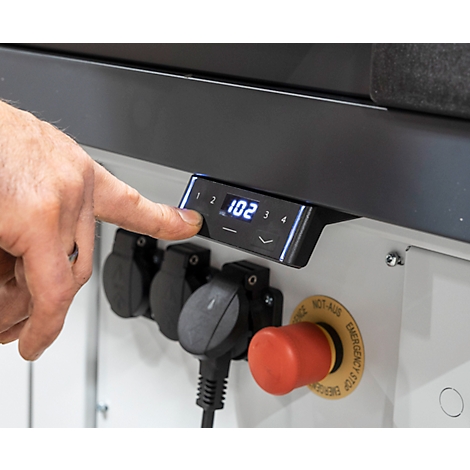 Close-up van een hand die een bedieningspaneel met een digitale display bedient. Hieronder bevinden zich stopcontacten en een rode noodstopknop.