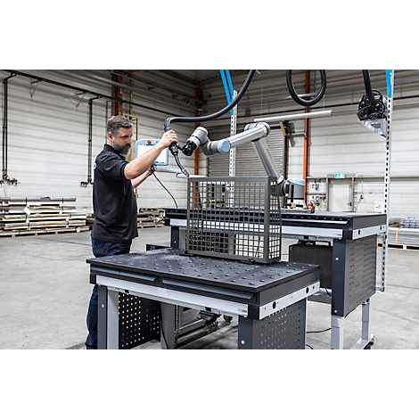 Een man bedient een robotarm die een metalen mand op een tafel inspecteert. Fabriek op de achtergrond.