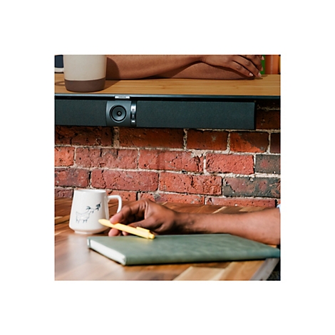 Nahaufnahme: Eine Hand hält einen gelben Stift über einem grünen Notizbuch. Eine Tasse und eine Soundbar sind zu sehen.