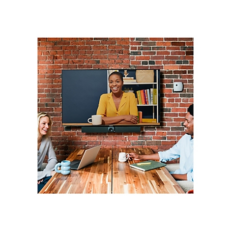 Eine Frau in Videokonferenz lächelt, während zwei Personen an einem Tisch sitzen. Der Hintergrund ist eine Backsteinmauer.