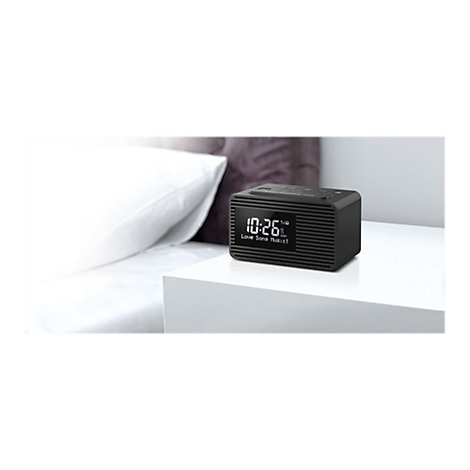 Ein schwarzer Radiowecker auf einem Nachttisch, im Hintergrund ein Bett. Auf dem Display steht die Uhrzeit 10:26 und der Radiosender.
